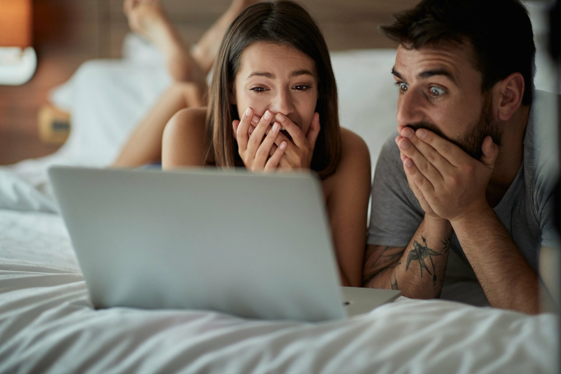 un homme et une femme allongés dans un lit sont stupéfaits en regardant une vidéo sur un ordinateur portable
