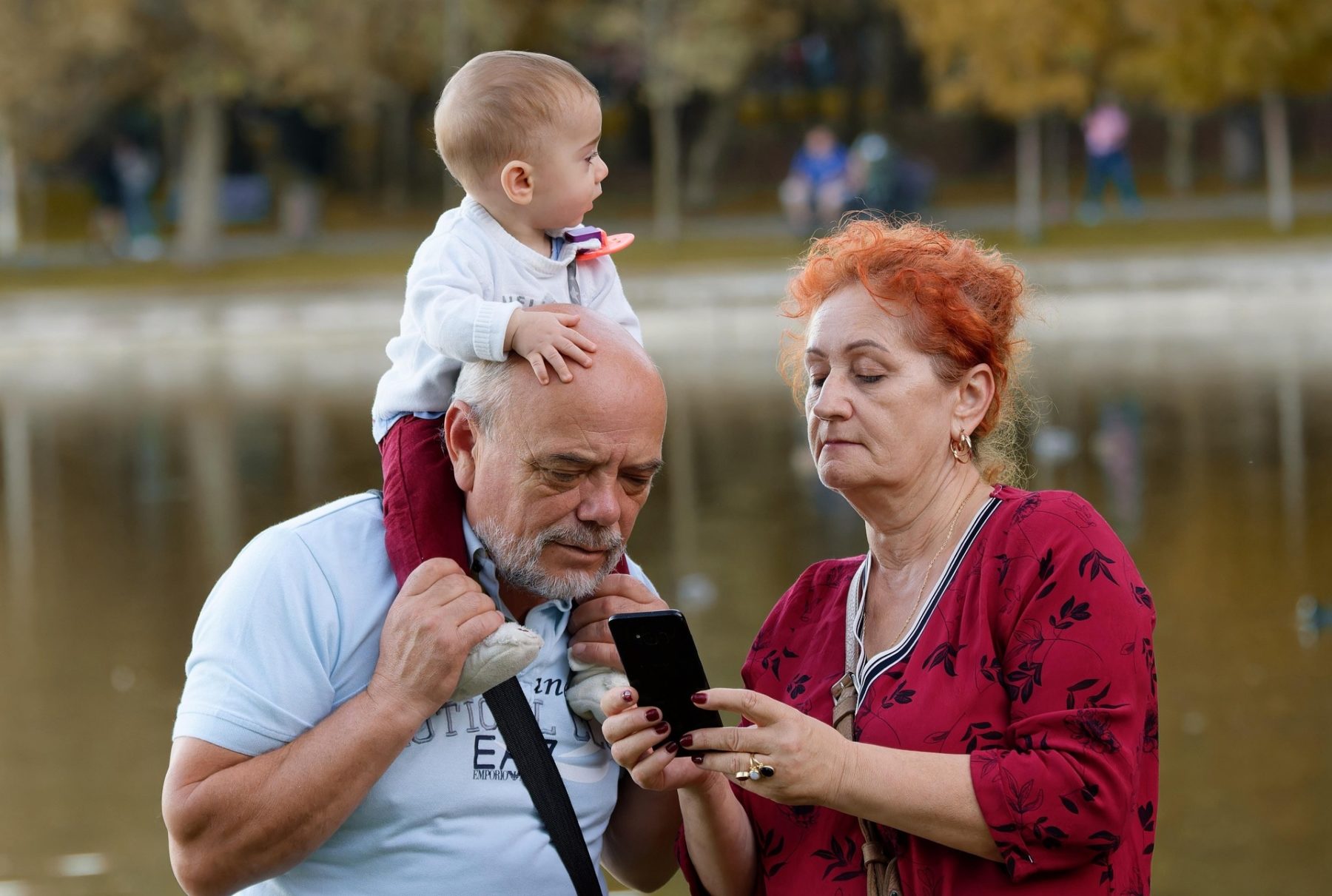 un grand père porte son petit-fils sur les épaules et regarde l'écran du smartphone que tient sa femme à côté de lui. Ils sont près d'un lac dans la nature.