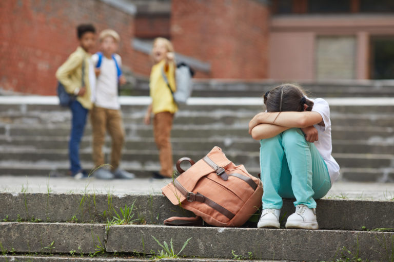 Une fille assise sur des marches pleure, ses camarades au second plan semble se moquer d'elle